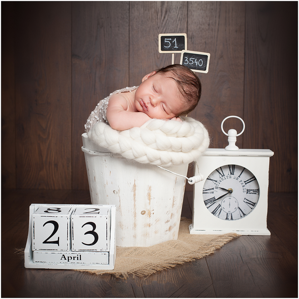 Baby Fotoshooting Duesseldorf: Neugeborenes Baby schläft in einem Holzeimer in einem Wollschal. Die Daten seiner Geburt sind erkennbar durch eine Uhr, einen Kalender und kleine Tafeln.